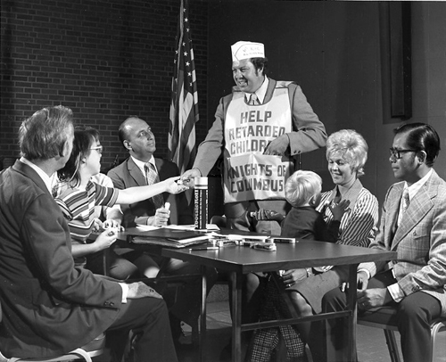 Parents sit around a table with two children. A man stands in an apron that reads 