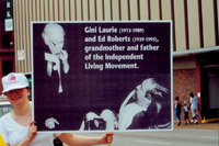 A woman holds up a sign that depicts Gini Laurie and Ed Roberts and includes the caption 'Gini Laurie and Ed Roberts, grandmother and father of the Independent Living Movement.'